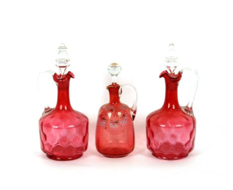 A pair of cranberry dimpled glass claret jugs,&nbsp;having clear glass handles and stoppers; and another with blue floral ena