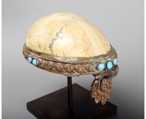 A Tibetan skull libation cup with embossed copper mounts and turquoise coloured beads, 18.5cm long, on a stand. (2)