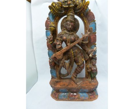 An Indian carved wood and painted temple panel of the Goddess Saraswati playing a sitar, on lotus flower base and beneath an 