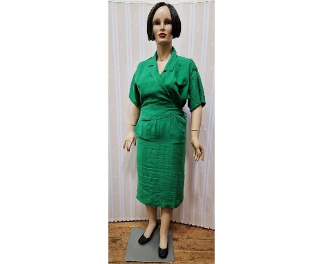 A vintage female mannequin, full figure, painted face and short wig, wearing a green linen wrap dress, on a metal stand, can 