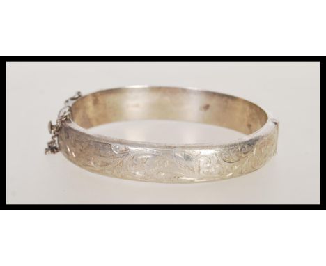 A hallmarked silver bangle bracelet having engraved scrolled detailing, with a hinged opening and safety chain. Hallmarked Bi