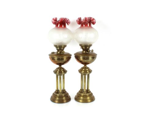 A pair of brass table oil lamps, supported on three Corinthian columns, ruby tinted flared shades, 73cm high