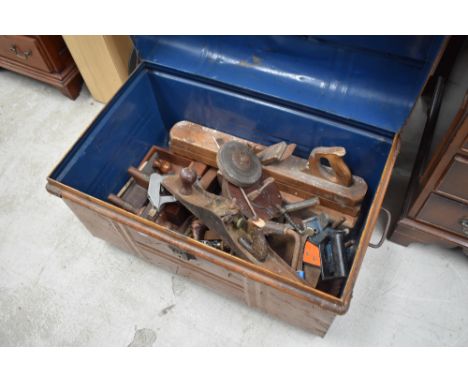 A vintage tin trunk with wood grain decoration containing a selection of hand tools including planes.