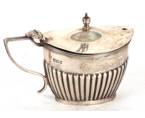 A late Victorian silver oval shaped mustard with a reeded rim, hinged lid with an urn finial, the body part fluted decorated,