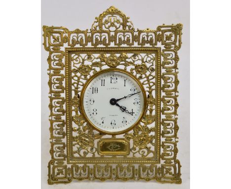 A late 19th century brass cased easel back timepiece, the ornate scrolling frame centred with a white enamel dial set with Ar