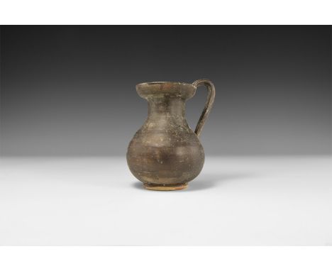 5th-3rd century BC. A ceramic blackware jug with discoid base, bulbous body, waisted neck, flared mouth, strap handle to the 