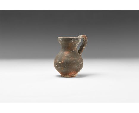 5th-3rd century BC. A ceramic blackware juglet with piriform body, discoid base, flared rim, flat-section strap handle to the