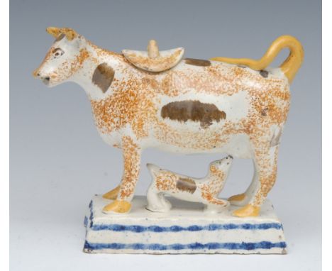 A Prattware cow creamer and cover, standing four square, sponged in ochre, tan patch markings, yellow horns and tail, with su