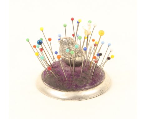 A silver pincushion in the form of a chick emerging from an egg, seated on a domed purple cushion within a circular silver fr
