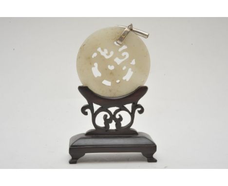 A Chinese carved hard stone pendant with white metal mounts, possibly jade and carved wooden stand. Diameter: 5.5cm approxima