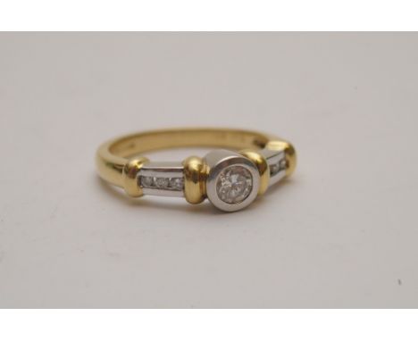 A ladies 18ct dual tone gold ring of modern design bezel set with a brilliant cut diamond solitaire, the shoulders channel se