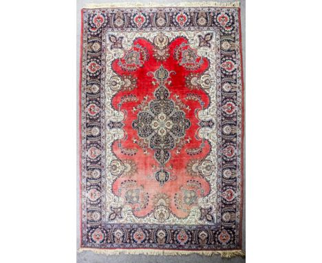 A Kirman carpet woven in colours with a bold shaped central medallion and floral palmettes on a plain red ground, conforming 