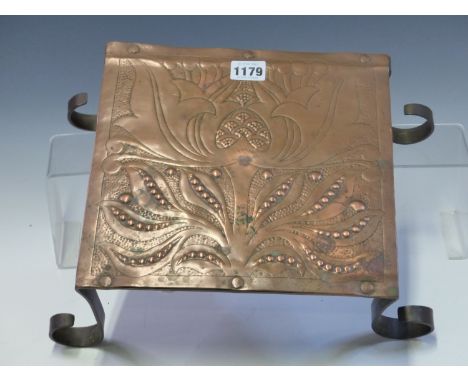 AN ARTS AND CRAFTS COPPER TRIVET, POSSIBLY BY C R ASHBEE, THE TOP WORKED WITH A FLORAL DESIGN ABOVE STRAP LEGS ON CURLED FEET