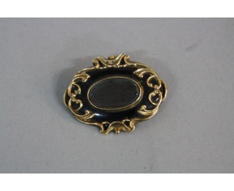 A MID VICTORIAN MOURNING BROOCH, black enamel with gold scroll design centring on a vacant glass compartment, measuring appro