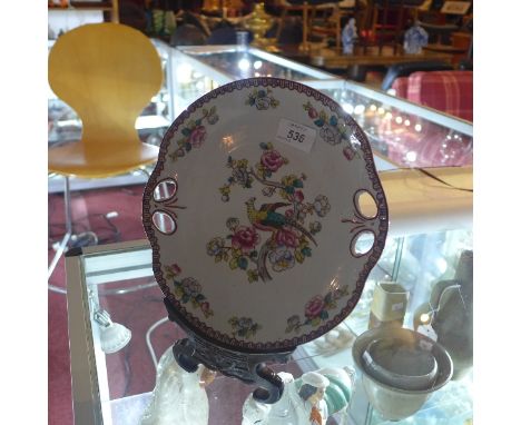 A ceramic plate having floral border and exotic bird with foliage to centre on a hard wood stand. A gold anchor mark to back.