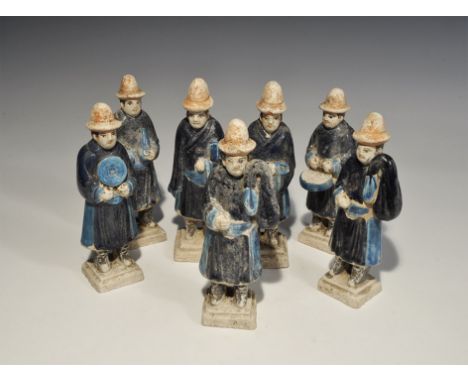 "20th century AD. A mixed group of glazed ceramic figurines, each in a blue robe and orange brimmed hat, holding a pan, bag o
