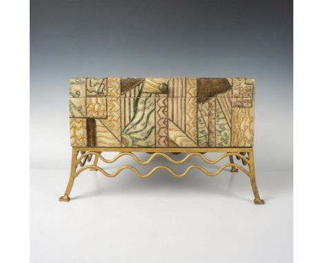 Raised abstract design colored in earth tones of browns and greens set on gilded metal stand. Round pull opens lid to felt li