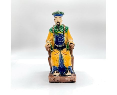 Glazed ceramic figure of a man in traditional yellow tunic shirt with blue accents, green accents and hat sitting on a chair.
