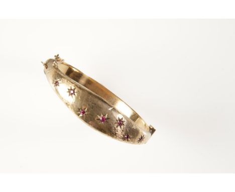 A 9CT YELLOW GOLD AND RUBY BANGLE, hinged, the tapering top section inset within engraved stars, with seven graduated round-c