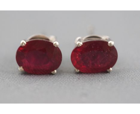 A pair of white metal single stone stud earrings. Each set with an oval faceted cut composite ruby (glass filled). Post and s