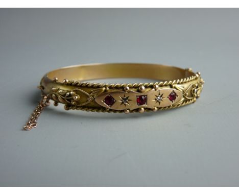 A NINE CARAT GOLD DIAMOND & RED STONE SET BANGLE with bead and rope decoration with floral shoulders and central cartouche of