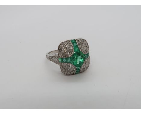 A platinum Art Deco style ring set with central oval cut emerald surrounded by calibre cut emeralds and round cut diamonds wi