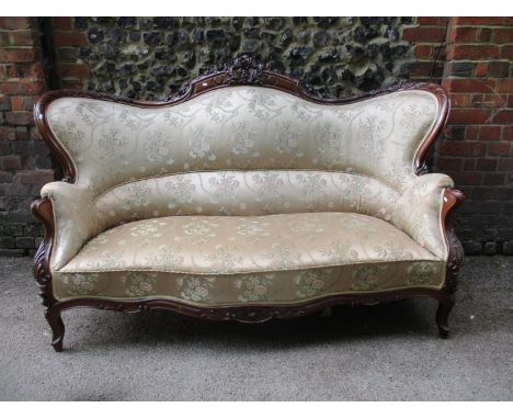 A late 19th century walnut framed sofa having a floral and foliage carved crest, curved wings and arms, upholstered in a beig