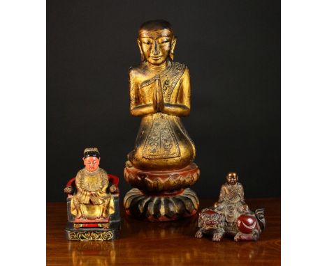 Three Far Eastern Polychromed Wooden Carvings: A Gilded Buddhistic Figure with hands held in prayer knelt on a lotus flower p