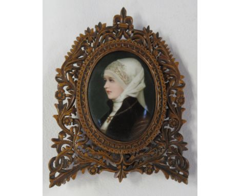 An oval porcelain plaque, painted with a portrait of a woman wearing a white headscarf, in a pierced, carved olive wood frame