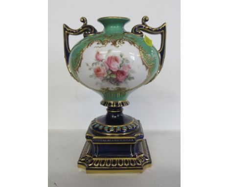 A Royal Worcester pedestal vase, decorated with a reserve of flower by Chare, to a green and gilt ground, with blue handles a