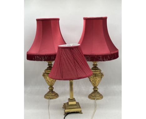 A pair of gilt lamps along with a brass column lamp, all with maroon shades.