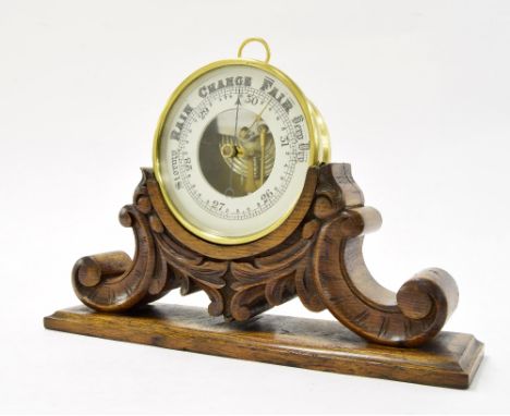 Brass drumhead aneroid desk barometer, the 4.75" chapter ring enclosing a skeletonised centre, upon a foliate carved oak stan