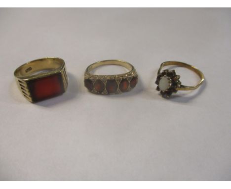 Three 9ct yellow gold gem set rings comprising a garnet five stone ring, an opal and garnet cluster ring, and a red stone pla