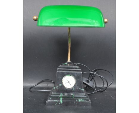 A 20th century converted bankers lamp with a green glass shade upon a stepped faux marble base with a clock to the centre. Me