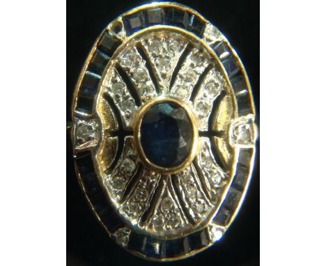 An Art Deco diamond and sapphire panel ring, central oval dark blue sapphire, surrounded by an open oval shield, encrusted wi