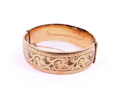 A yellow metal hinged bangle. Having engraved decoration and a clip-lock clasp with a safety chain. Marked '1/5 9ct Metal Cor