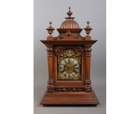 A German carved bracket clock, with fish scale detailing, reeded columns and turned finials. Featuring brass figural dial and