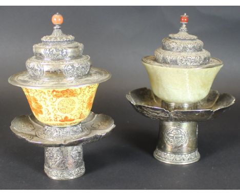 Two similar Tibetan white metal mounted offering cups on stands, one with a yellow ground porcelain cup, the other with a pal