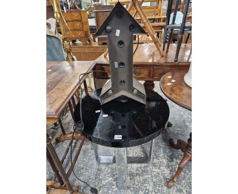 An electrically lit arrow sign together with a black circular table on chrome legwork