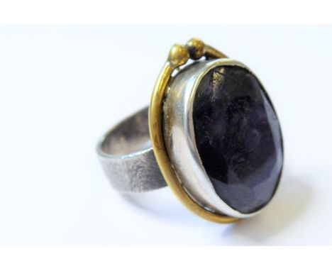 A bespoke silver ring; the textured ring with large applied central dark-blue hand-cut stone with a yellow-metal wire, ring s
