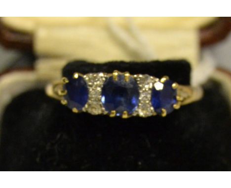 A diamond and sapphire ring, central cushion cut mid blue sapphire flanked by a smaller conforming stone either side, each di
