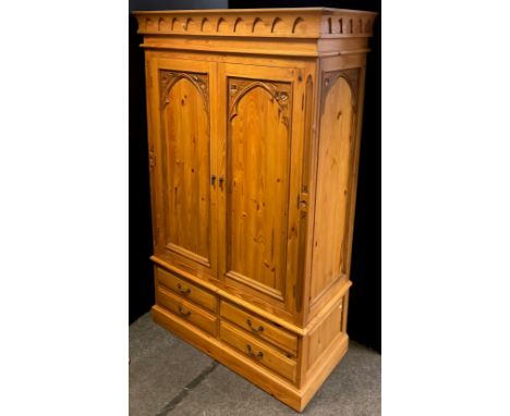 A Gothic style pine double wardrobe, having a pair of doors with Gothic arch carved panels, above four short drawers to base,