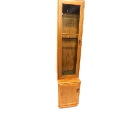 An Ercol light elm narrow cabinet, the top with a single glazed door with bevelled glass, the base with a door with a shaped 