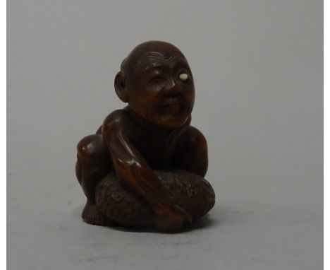 A Japanese wood netsuke of a blind beggar, 19th century, attempting to lift his distended scrotum, one eye with ivory inlay, 