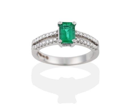 An 18 Carat White Gold Emerald and Diamond Ring, the emerald-cut emerald in a four claw setting, to tramline shoulders set wi