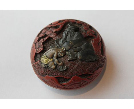 A small Japanese cinnabar circular manju shaped netsuke, the cover decorated with metal figures of devil kneeling before a 'g
