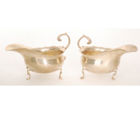 A pair of hallmarked silver sauce boats each raised on three pad feet below waved borders and flying scroll handles, London 1