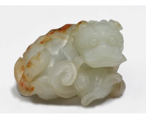 A CHINESE WHITE AND RUST JADE PEBBLE carved as a reclining dog of fo holding a tassel of a ball which rests beneath its paws,