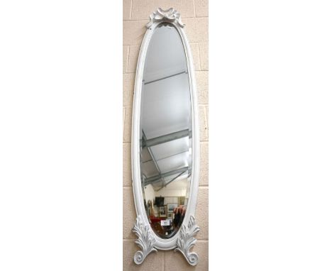 A narrow bevelled oval wall mirror in ribbon crested and foliate carved cream painted frame, 148 cm high x 36 cm wide