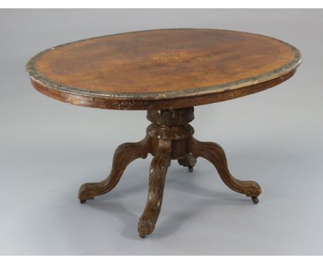 A late Victorian walnut tilt-top oval dining table with marquetry inlaid decoration &amp; carved edge, on foliate carved bulb